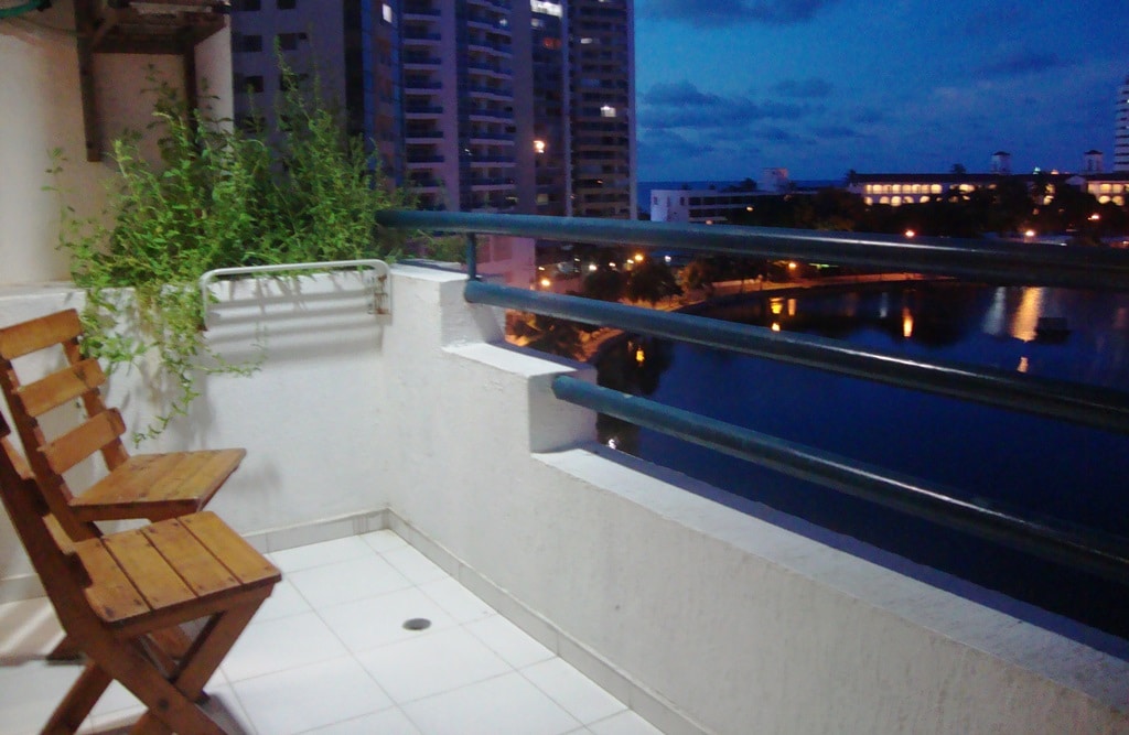 A balcony space features two wooden chairs positioned by a railing. A green plant is displayed in a planter beside the chairs. City lights shimmer over the water in the background, creating a serene evening atmosphere.