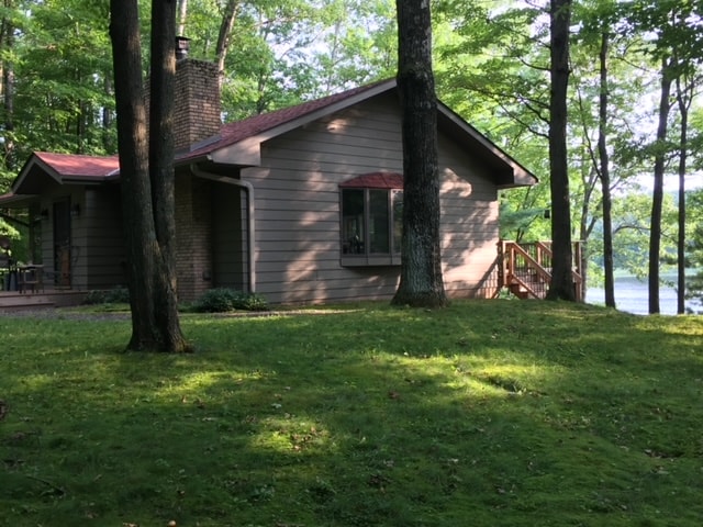A rustic cabin is nestled among trees with a serene lake visible in the background. The exterior features a warm color palette and a red metal roof. A porch with steps leads to a grassy area, inviting leisurely outdoor activities.