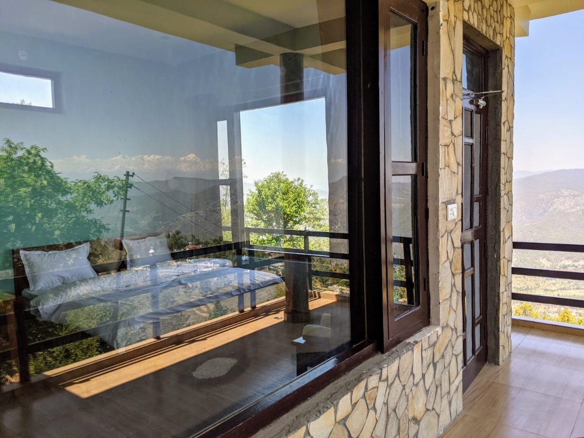 A glass-enclosed bedroom offers panoramic views of the surrounding landscape. The room features a spacious bed with crisp linens, while natural light floods in through large windows. The stone exterior provides a rustic charm, complementing the serene mountain backdrop.