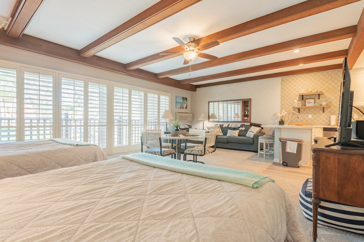 A spacious room features three queen-size beds, flanked by wooden beams on the ceiling. A comfortable seating area with a sofa and a small dining table is visible. Natural light floods in from large plantation-shuttered windows, enhancing the room’s airy ambiance.
