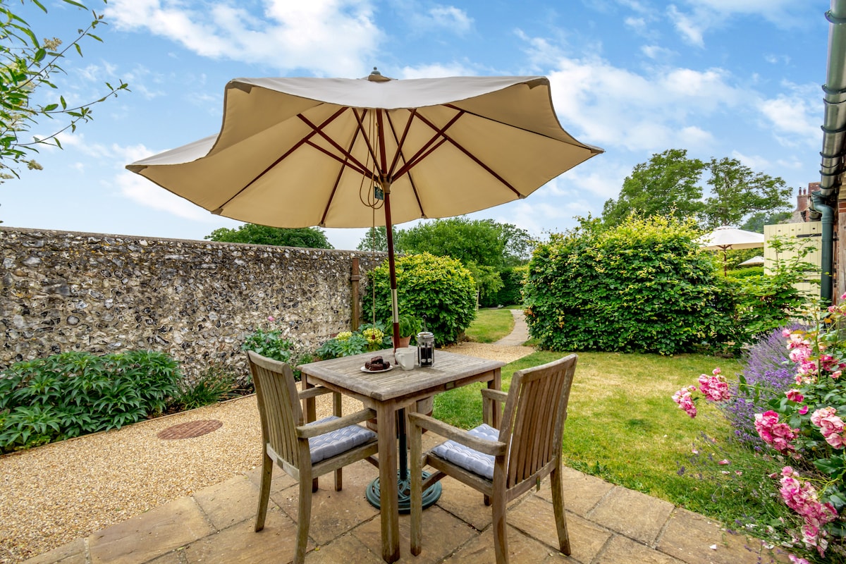 A quaint outdoor terrace features a wooden table surrounded by two chairs, shaded by a large umbrella. Lush greenery and colorful flowers accentuate the area, providing a peaceful setting for enjoying meals or relaxing outside.