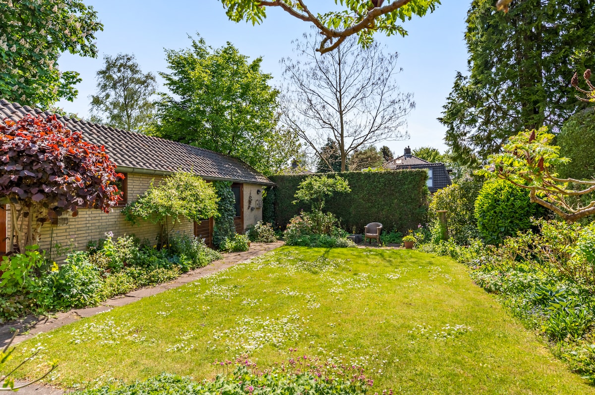 A sunny garden is showcased featuring well-maintained grass and flowerbeds. A stone pathway leads to a separate garden house nestled among greenery. Various trees and shrubs provide a natural perimeter, enhancing privacy and tranquility in the outdoor space.