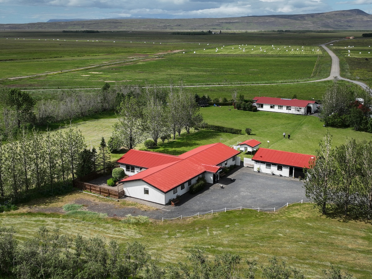 An aerial view showcases the guesthouse and surrounding grounds, featuring a traditional design with a red roof. The expansive green landscape includes lush fields and scattered trees, with additional buildings visible in the distance. The peaceful setting highlights the beauty of the Icelandic countryside.
