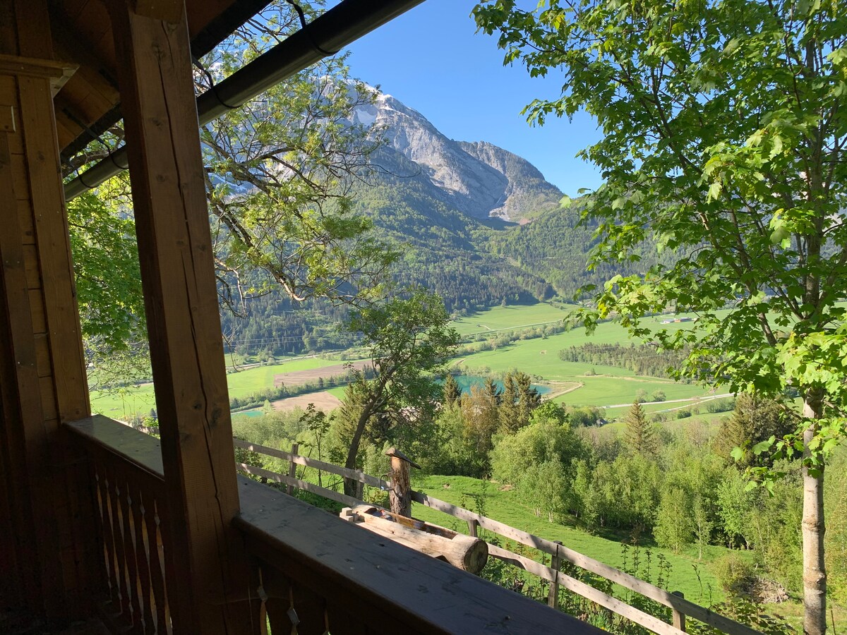 The image showcases a panoramic view from a wooden balcony, with lush green hills and trees in the foreground. A mountain range rises in the distance under a clear blue sky, emphasizing the natural beauty of the surrounding landscape.