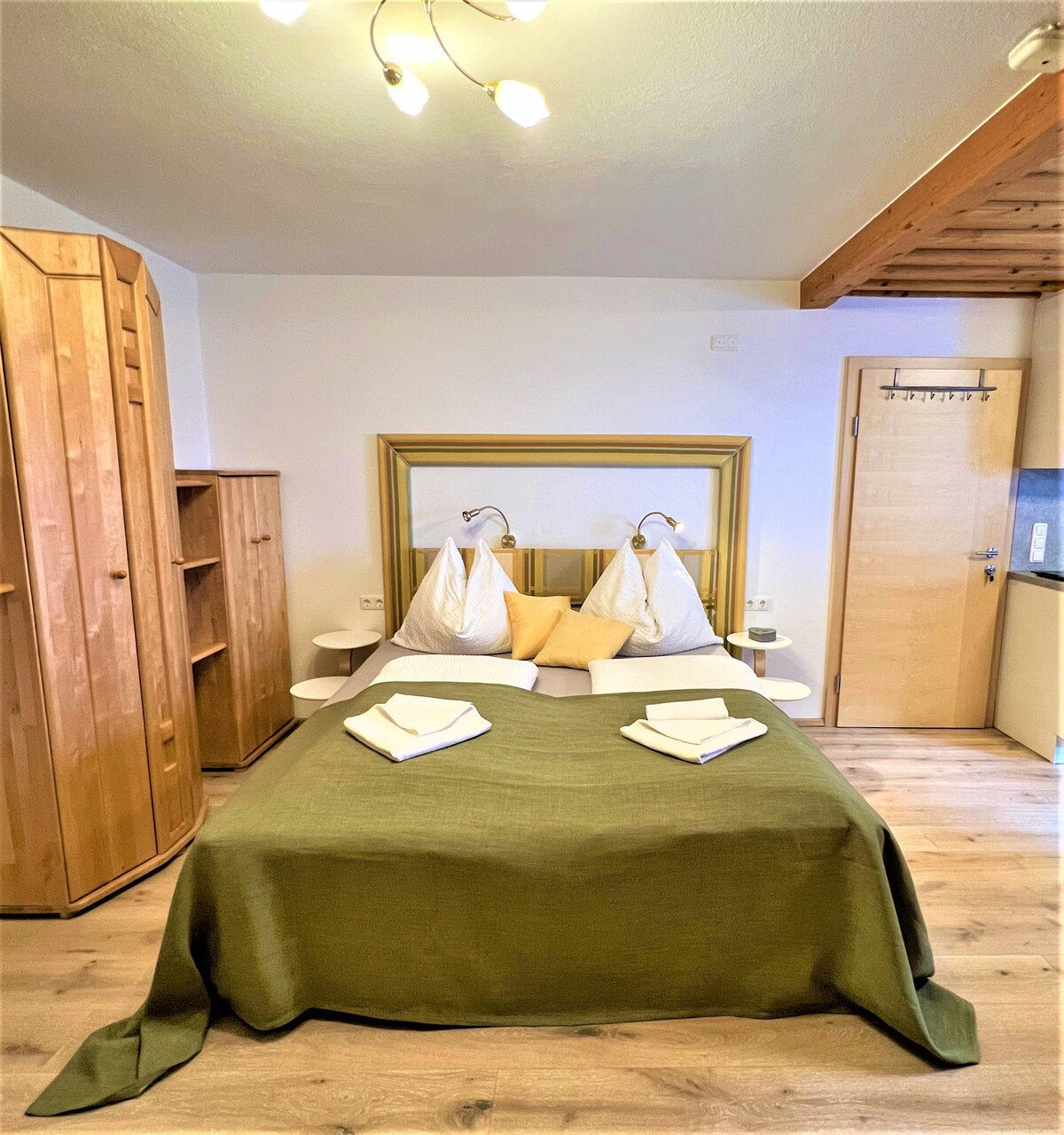 A cozy sleeping area is presented, featuring a spacious bed adorned with a green blanket. Two decorative pillows and freshly folded towels are arranged atop the bed. Wooden cabinetry offers storage, while soft lighting from a ceiling fixture enhances the ambiance.