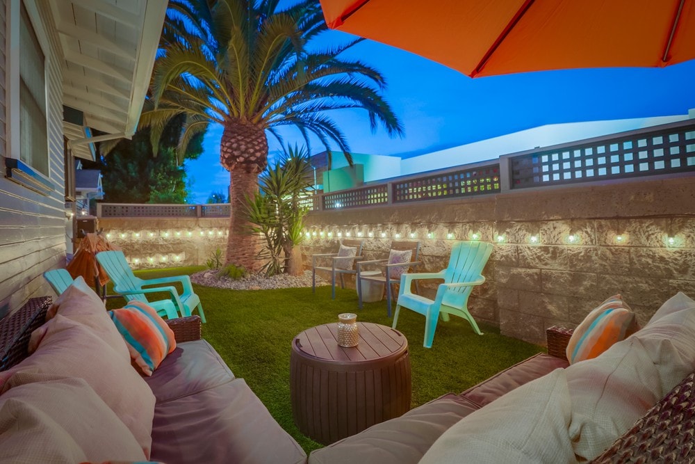 An inviting outdoor area is presented featuring a cozy sectional sofa with colorful cushions. Lounge chairs in light blue are positioned beside a palm tree, with soft lighting illuminating the space. An orange umbrella provides shade, creating a comfortable setting for relaxation.