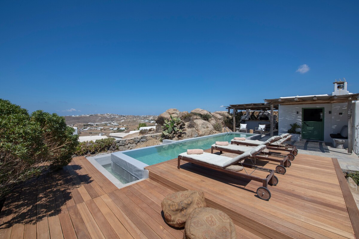 A sunlit outdoor area features a private swimming pool surrounded by wooden decking. Sun loungers with soft cushions are positioned for relaxation. Lush greenery provides a natural border, and the distant view of white-washed buildings highlights the serene landscape.
