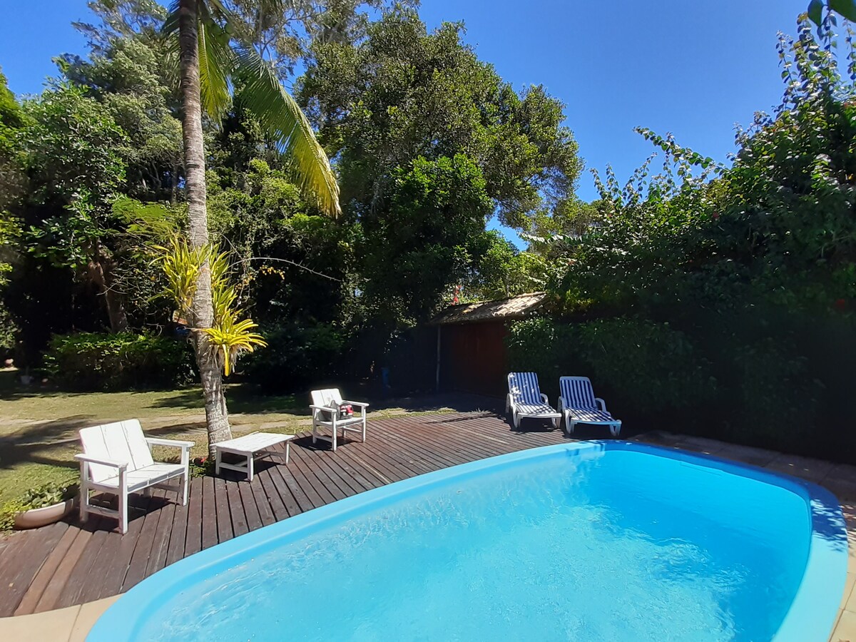 A private swimming pool is surrounded by a wooden deck, with several lounge chairs positioned nearby. Lush greenery and tall trees provide a natural backdrop, enhancing the outdoor leisure area. The clear blue sky complements the tranquil atmosphere.