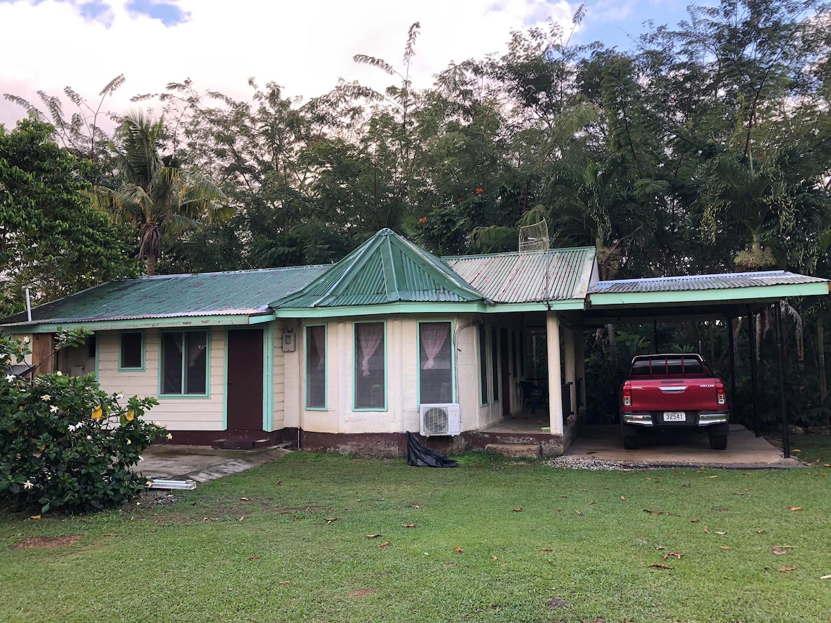 The exterior of the house features a green roof and light-colored walls with large windows. A covered parking space accommodates a vehicle, while the surrounding garden is lush with tropical greenery. The property is set within a spacious, grassy area.