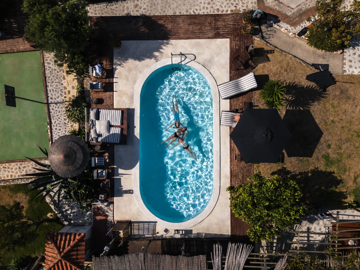 An aerial view captures a rectangular swimming pool surrounded by lush greenery and comfortable lounge chairs. The pool's clear water reflects sunlight, creating a serene atmosphere. Nearby, an outdoor dining area and a shaded seating space under a black umbrella are visible.