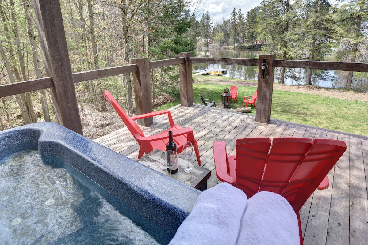 A private outdoor area features a hot tub next to a wooden deck with two red chairs. A view of the lake and surrounding greenery is visible. A small table holds a bottle of wine and two glasses, inviting relaxation in a serene setting.