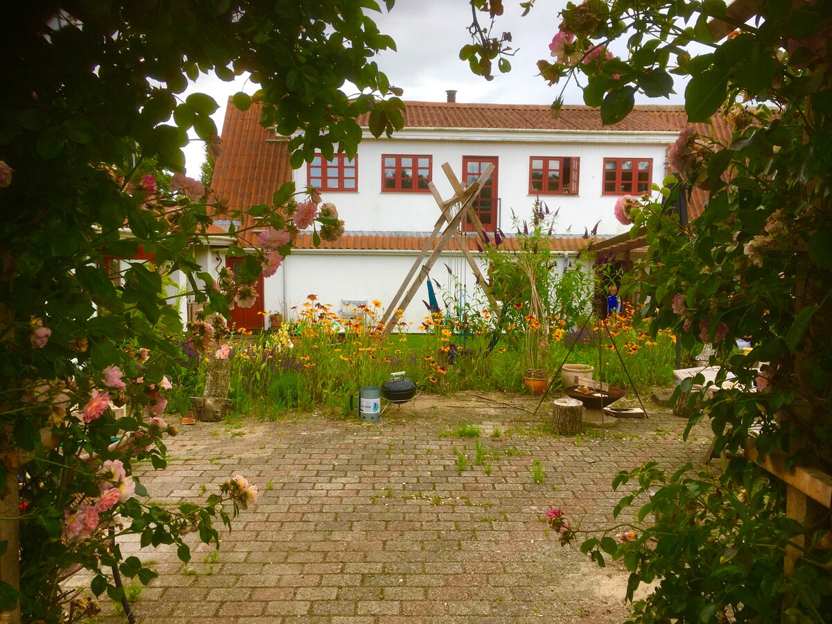 A gravel path leads into a charming garden filled with vibrant flowers and greenery. The two-story building is visible in the background, featuring red-framed windows and rustic architecture. Outdoor seating and wooden accents are arranged throughout the space, creating a welcoming atmosphere.