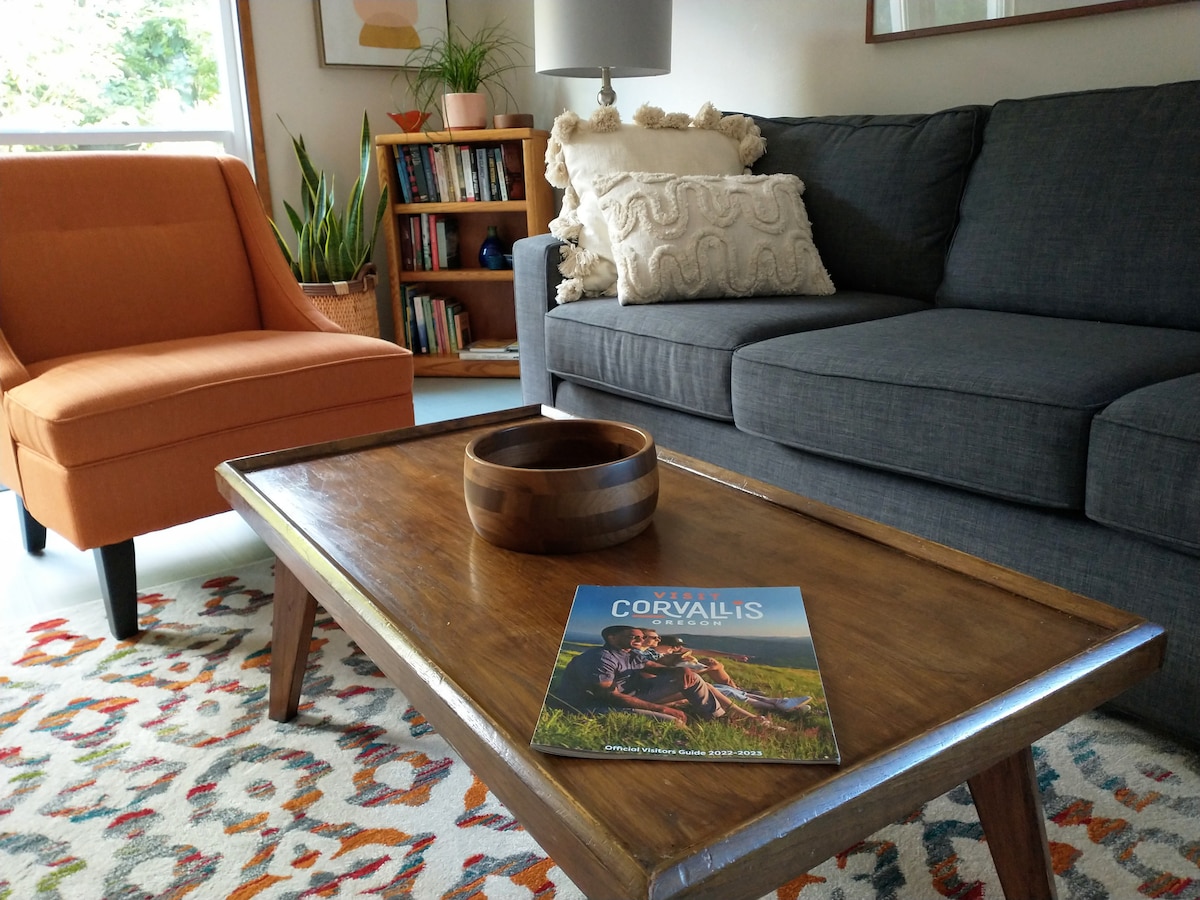 A cozy living area is displayed, featuring a gray sofa and an orange accent chair. A wooden coffee table sits in front, topped with a magazine. The space is enhanced by greenery and a bookshelf in the background, allowing for natural light to fill the room.