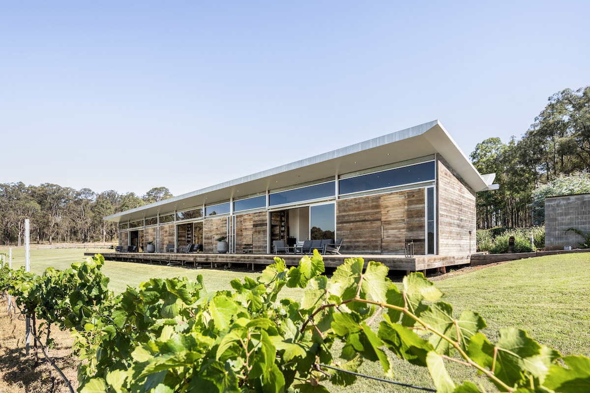 The modern structure features a sleek design with large glass panels that open up to expansive views of the surrounding vineyard. A spacious wooden deck is highlighted, providing an inviting outdoor area that seamlessly integrates with the lush green landscape.
