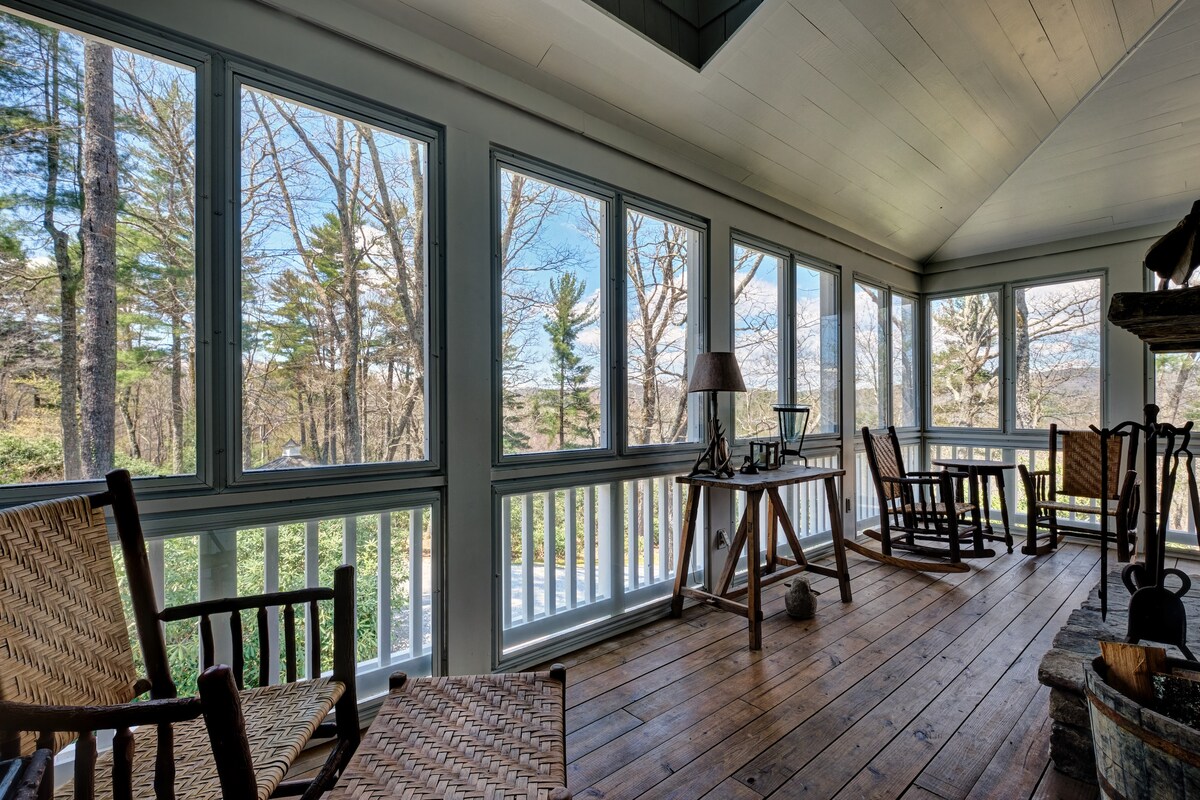 A spacious screened porch features large windows that provide a view of the surrounding trees and mountains. Multiple wooden rocking chairs and a small table are arranged on the warm-toned wooden floor, creating an inviting space for relaxation.