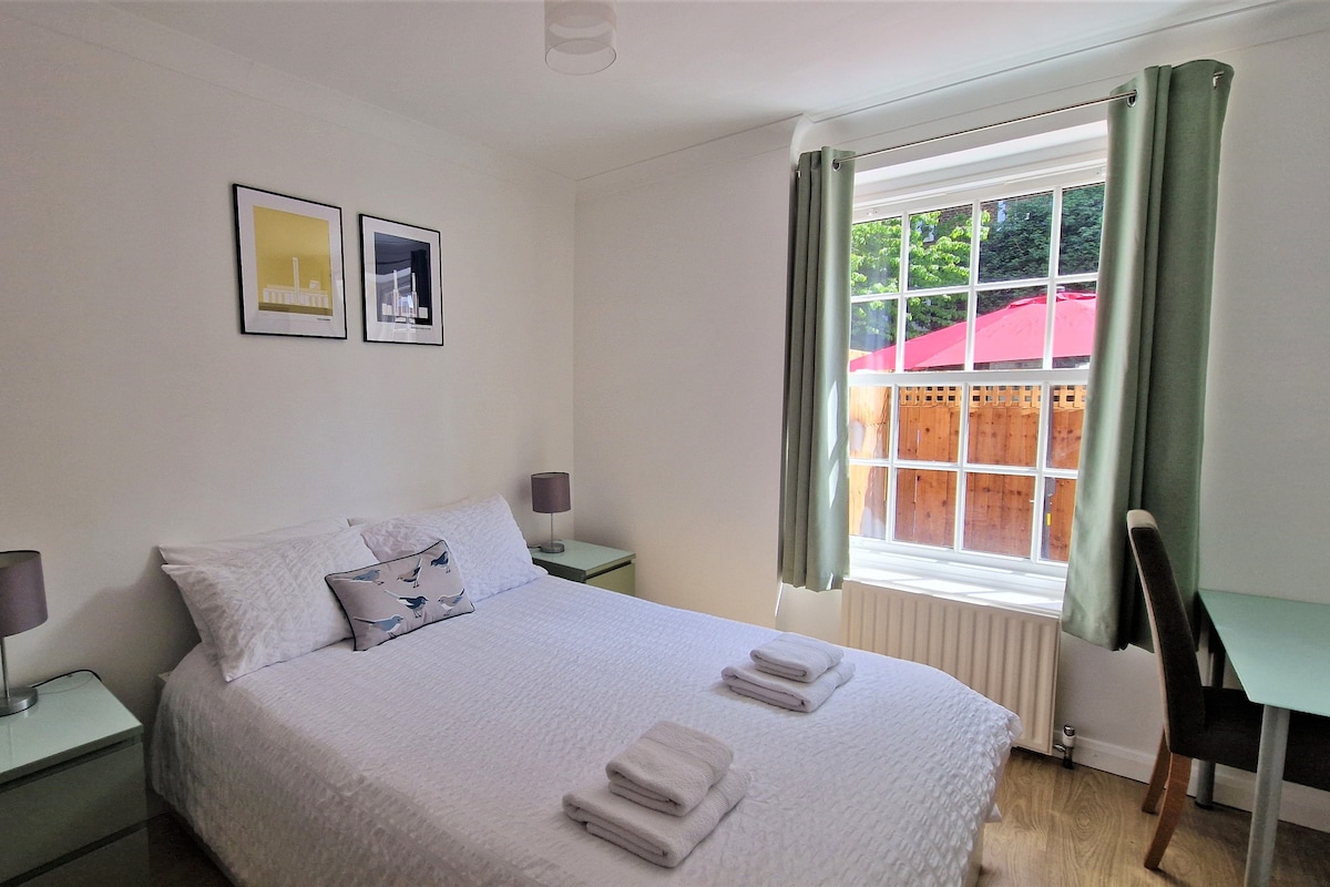 A bright double room features a spacious bed dressed in white linens, with neatly folded towels placed at the foot. Artwork adorns the wall above the bed, complementing the natural light that filters through the large window, which looks out onto a garden area.