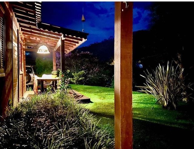The wooden house is illuminated by soft lighting, with a covered porch extending to a well-manicured lawn. Lush greenery surrounds the area, and a rustic dining table is visible on the porch, inviting relaxation in the evening atmosphere.