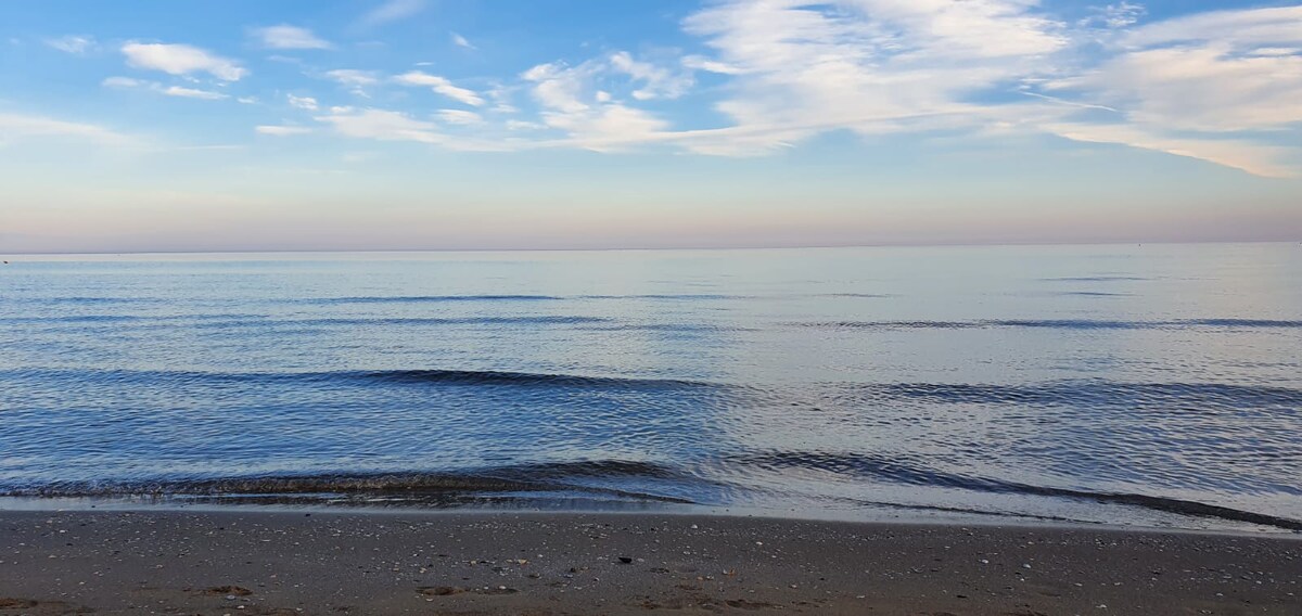 The image captures a serene beach landscape with gentle waves lapping at the shore. The calm sea reflects a gradient of blues and pinks under a clear sky, creating a tranquil atmosphere. Soft sand is visible in the foreground, dotted with small pebbles.