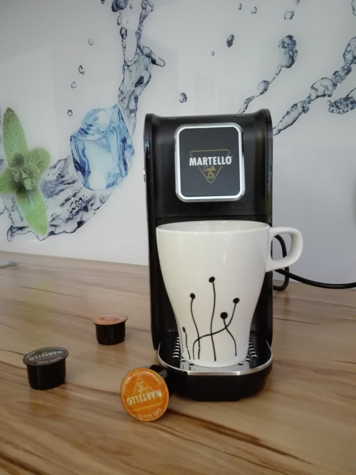 A coffee machine is positioned on a wooden countertop, with a white mug featuring a modern design placed underneath. Two coffee capsules in bronze and black are nearby, and a water-related graphic decorates the wall in the background.