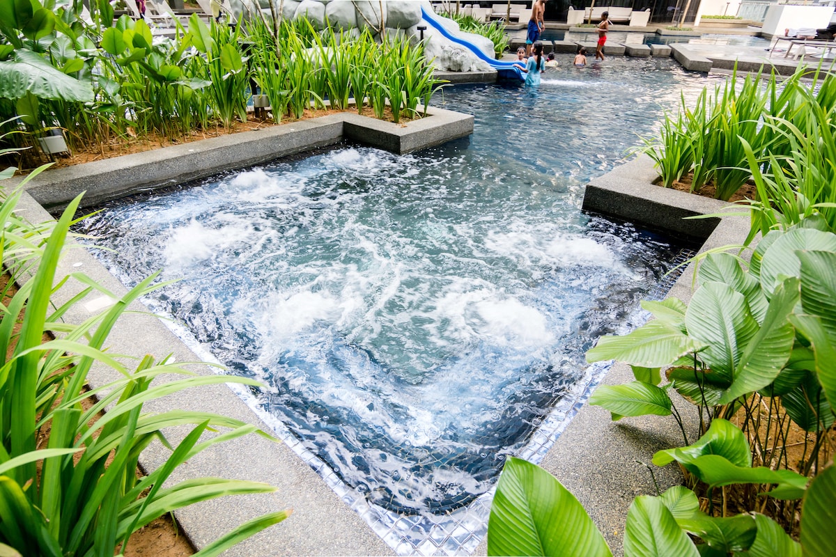 A refreshing swimming pool area is surrounded by lush green plants and modern stone pathways. Gentle waves ripple across the water's surface, creating a serene atmosphere. In the distance, children can be seen enjoying the water features, adding a playful touch to the setting.