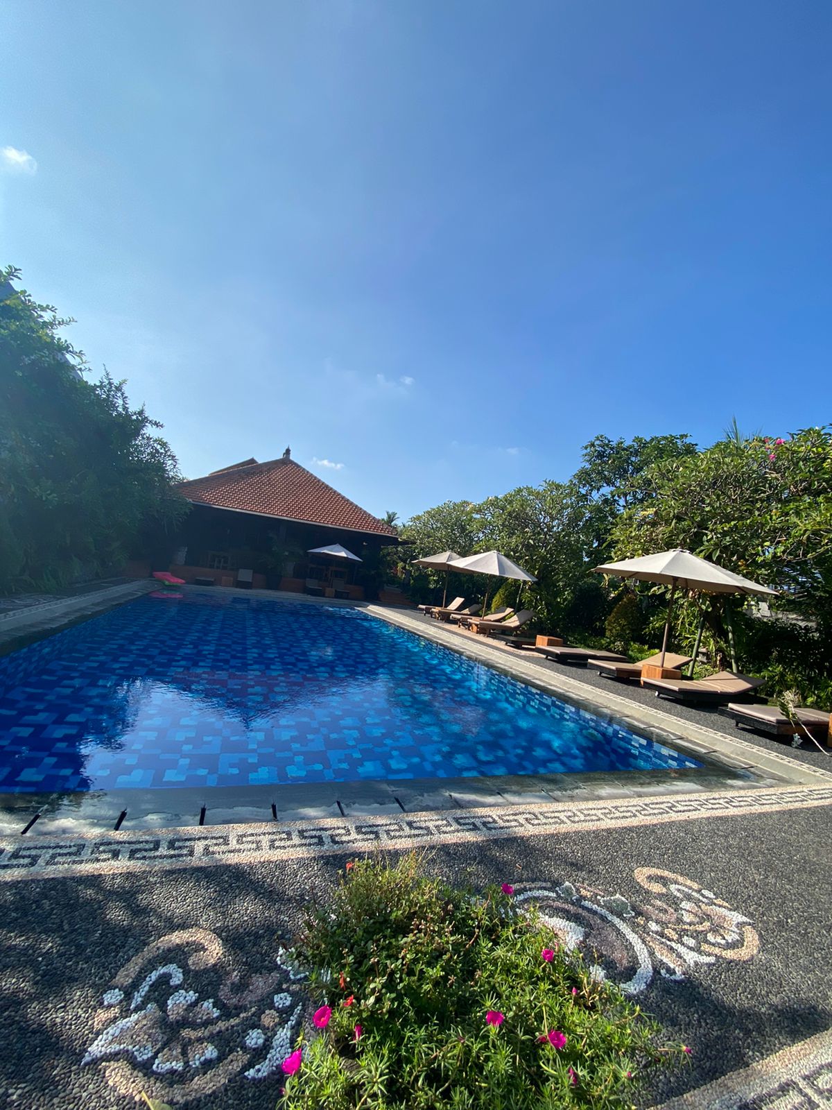 A large swimming pool features a vibrant blue mosaic pattern, surrounded by lush greenery. Comfortable lounge chairs are positioned near the water, complemented by umbrellas for shade. The setting includes a traditional structure in the background, adding to the serene atmosphere.