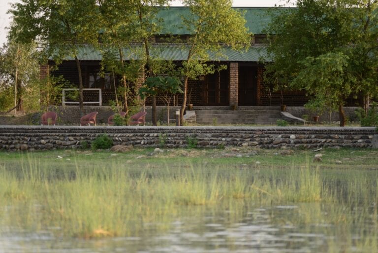 A calm view of a rustic building surrounded by lush greenery. A few chairs are positioned along a stone pathway, leading to a set of steps that access the entrance. The structure features a sloped roof with a green hue, complemented by the tranquil waters nearby.