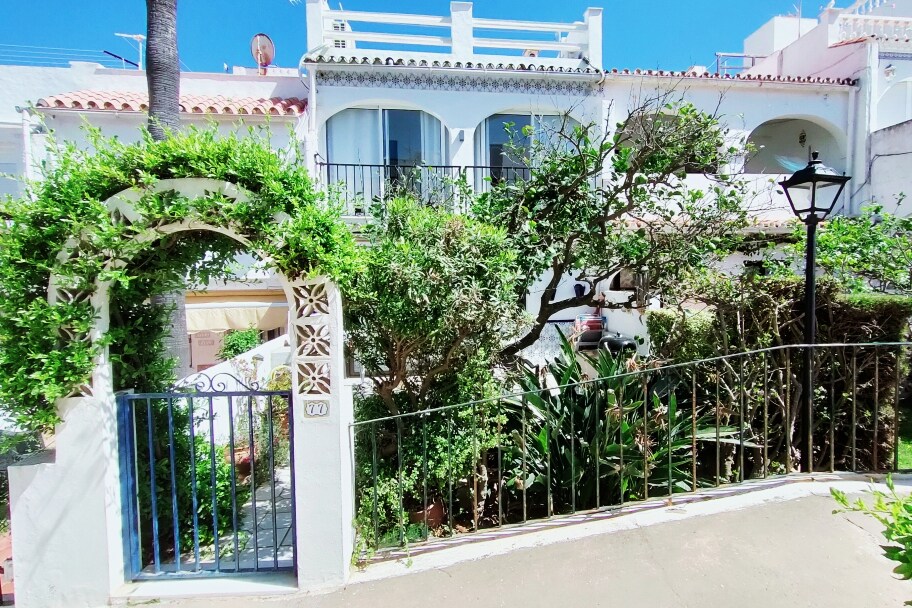 The entrance of the property is framed by a lush green archway, leading to a charming garden filled with various plants. A decorative gate opens to the path, alongside a lamp post that enhances the inviting atmosphere of the residential area.