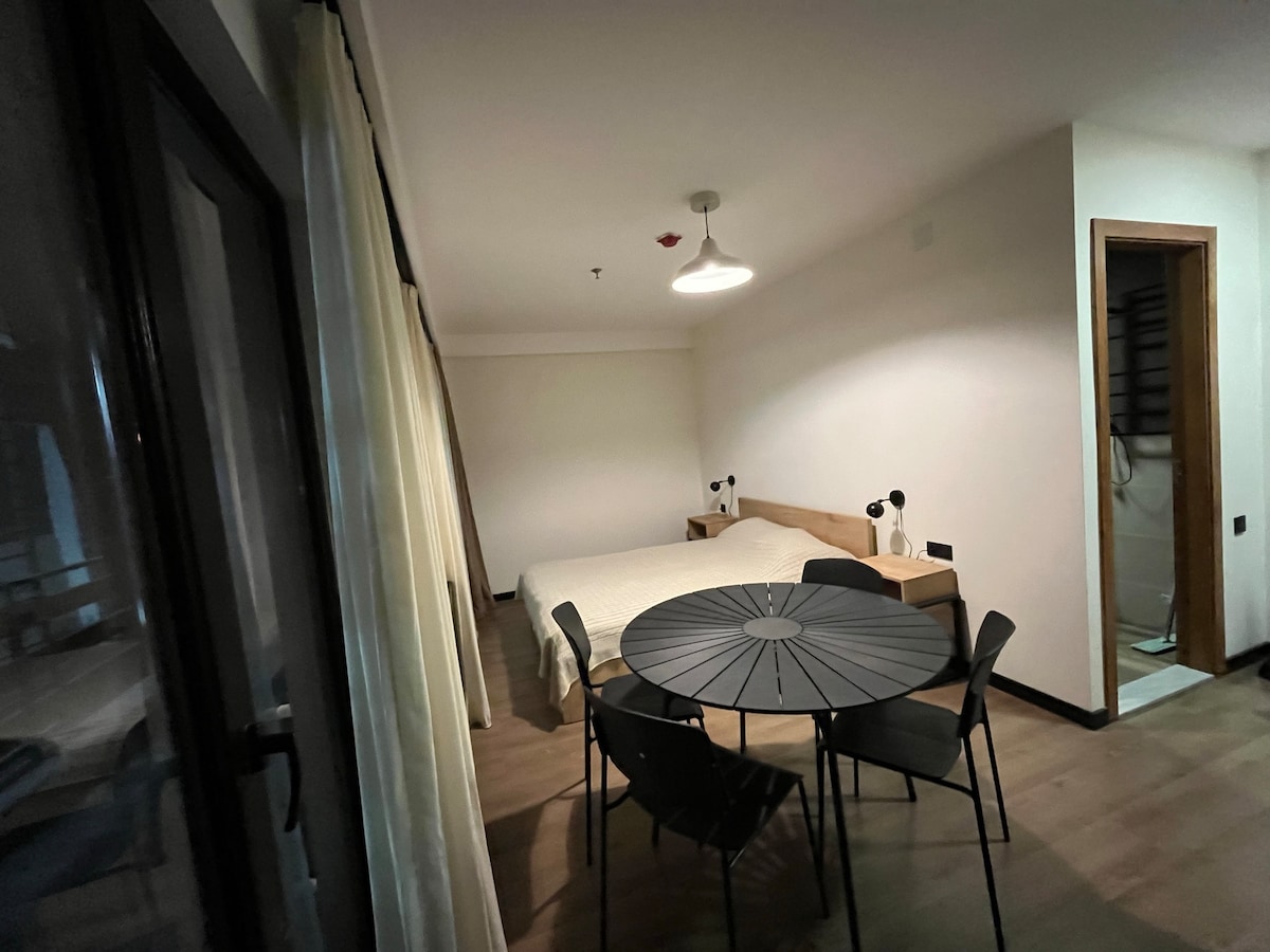 A spacious room features a large bed covered with a light blanket and a wooden bedside table on either side. A round dining table with several chairs occupies the center of the space. Natural light enters through sheer curtains, and a bathroom door is visible.