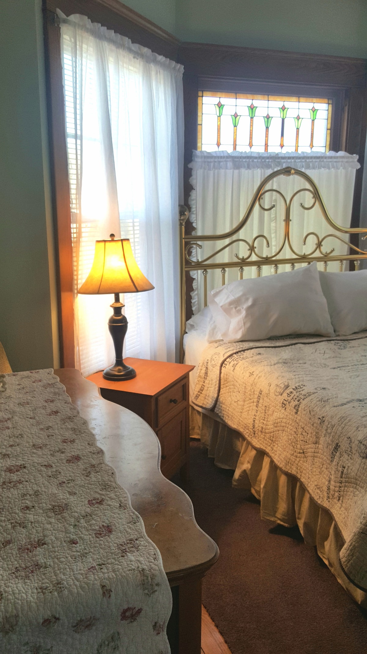 A bright and airy bedroom features a decorative brass bed with white bedding, complemented by a bedside lamp on a wooden table. Natural light filters through sheer curtains, highlighting a stained glass window above the bed. A dresser with a floral runner completes the room's aesthetic.