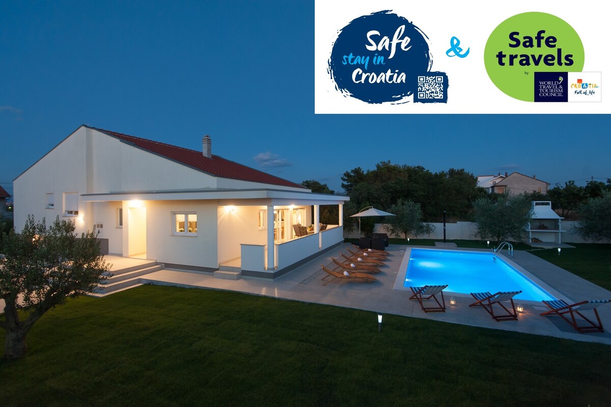 The exterior of a spacious villa is shown at dusk, featuring a swimming pool with clear water. Surrounding the pool are sun-loungers and a well-maintained lawn. An outdoor dining area is visible, along with a shaded spot provided by an umbrella.