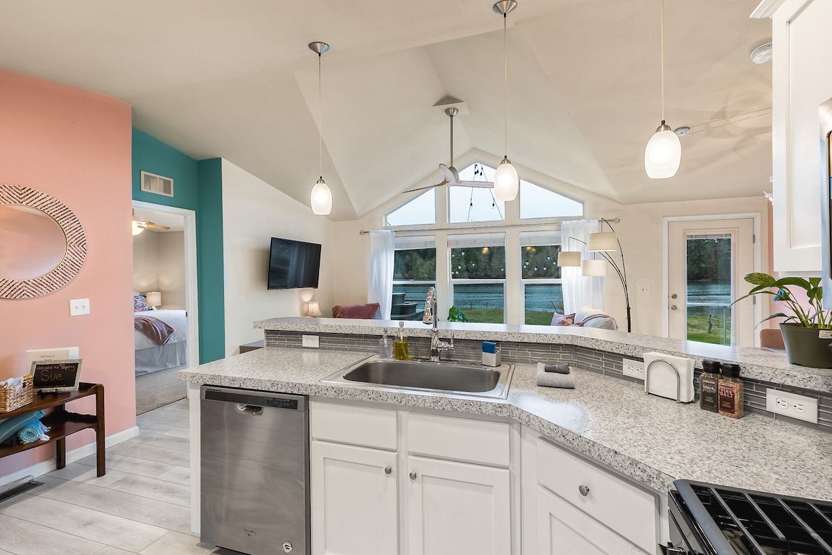 A bright kitchen area features light-colored cabinetry and a spacious countertop with a stainless steel sink. Modern light fixtures hang from the ceiling, and large windows offer a view of the water outside. A subtle color palette adds to the airy ambiance of the space.