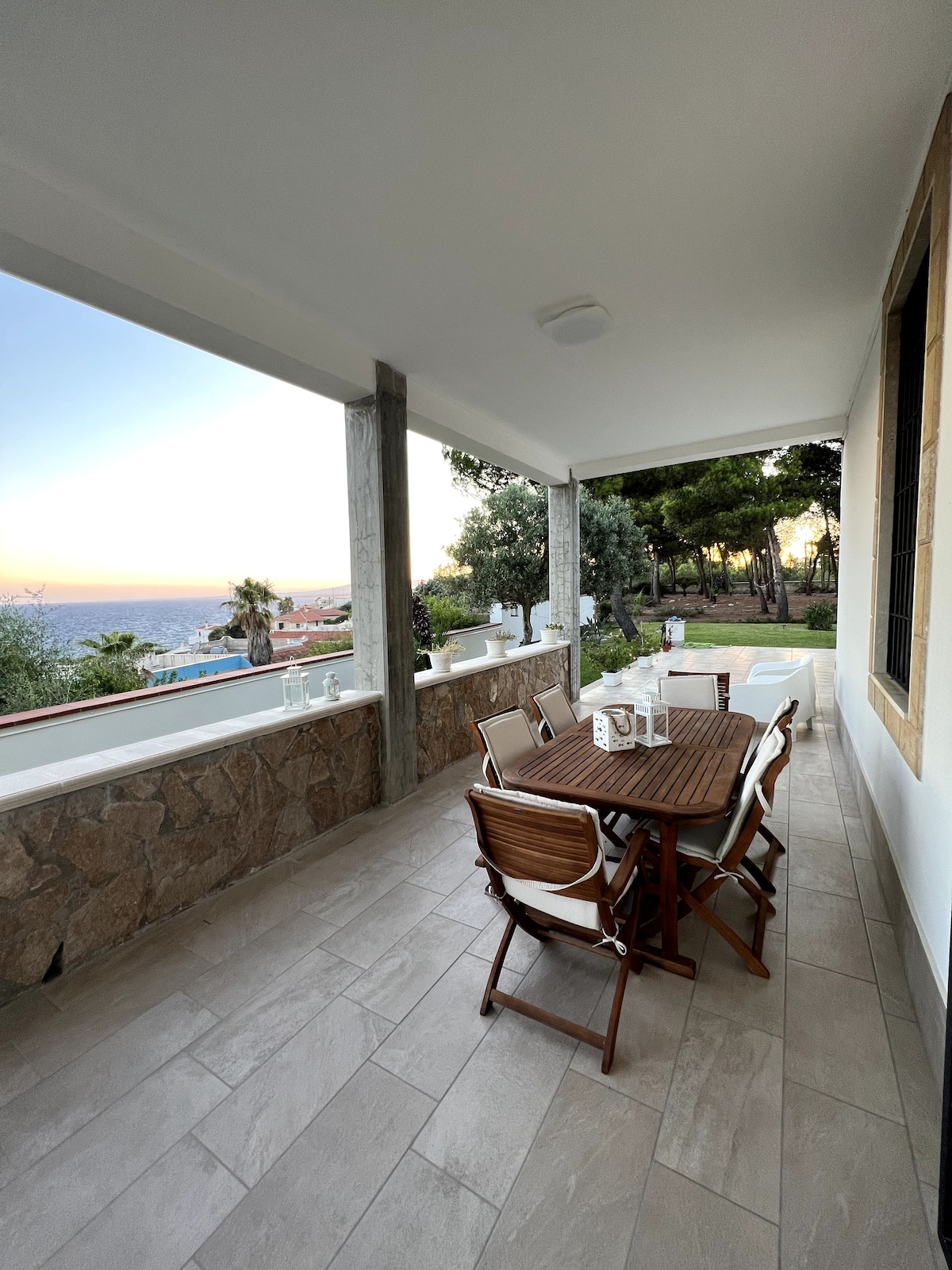 An outdoor terrace is presented with a classic wooden table surrounded by folding chairs. The scenic view includes a glimpse of the sea, framed by tall trees and greenery. Soft light from the setting sun enhances the relaxing atmosphere.