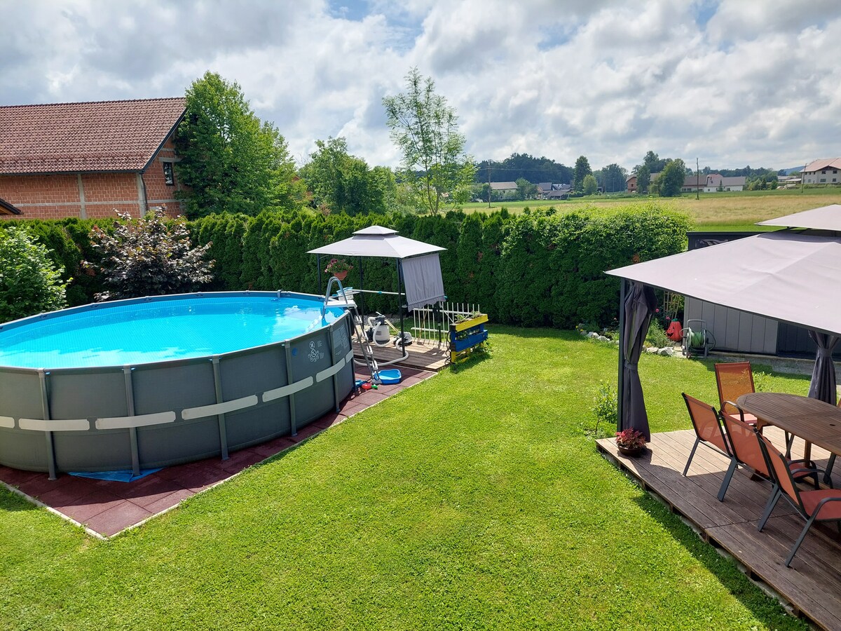 A spacious garden is featured, highlighting a round above-ground swimming pool surrounded by a grassy area. Several shaded seating sections are visible, equipped with tables and chairs. Lush green hedges border the garden, complementing the peaceful outdoor setting.