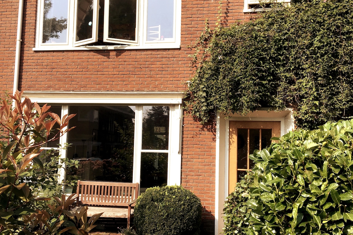 The exterior view features a brick facade adorned with creeping green vines. Large windows provide glimpses into the inviting interior. A wooden bench is positioned on the porch, surrounded by neatly trimmed hedges and lush greenery.