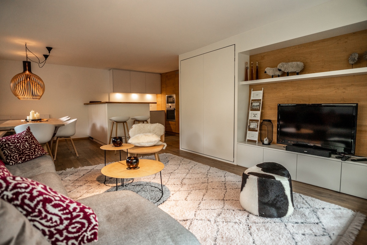 A modern living area features a spacious sofa adorned with patterned cushions and a soft area rug. A wooden shelf displays decorative items, while a flat-screen TV is mounted nearby. A dining table with white chairs and a unique pendant light adds warmth to the room.