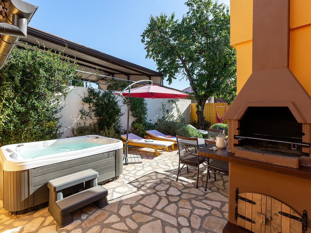 A private terrace is equipped with a whirlpool, sun loungers, and a shaded dining area. A barbecue facility is positioned next to the dining set. Lush greenery surrounds the space, providing a tranquil outdoor setting.
