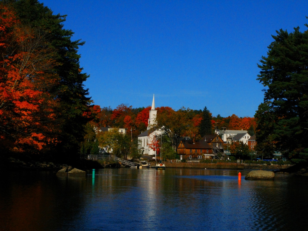 The image captures a serene view of a river reflecting brilliant autumn colors. A church steeple rises prominently against a backdrop of orange and red foliage, while charming buildings line the riverbank. The blue sky and calm water enhance the peaceful atmosphere.