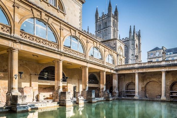 The historic baths feature classical columns and expansive arched windows that allow natural light to reflect off the tranquil water. A backdrop of intricate stone architecture and a tall cathedral spire adds to the scene, showcasing the beauty of the surroundings.
