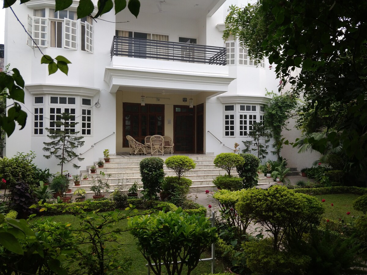 A two-story villa is framed by lush greenery and colorful flowerbeds. Steps lead up to a spacious entrance, featuring a covered patio furnished with wicker chairs. Large windows brighten the facade, creating a welcoming atmosphere amid well-maintained gardens.