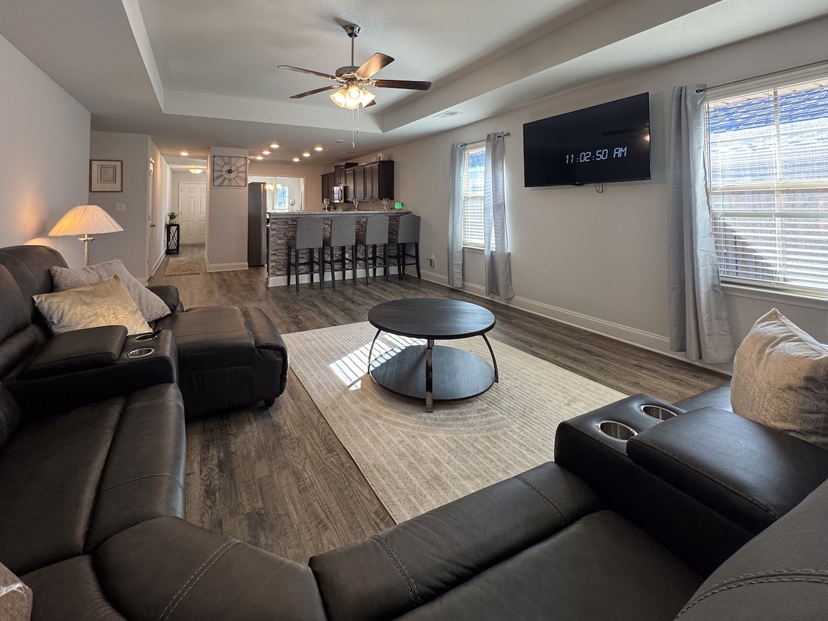 A spacious living area is featured, highlighting a reclining couch and a round coffee table in the center. A large flat-screen TV is mounted on the wall, and natural light enters through the window. Barstools line the kitchen island in the background.