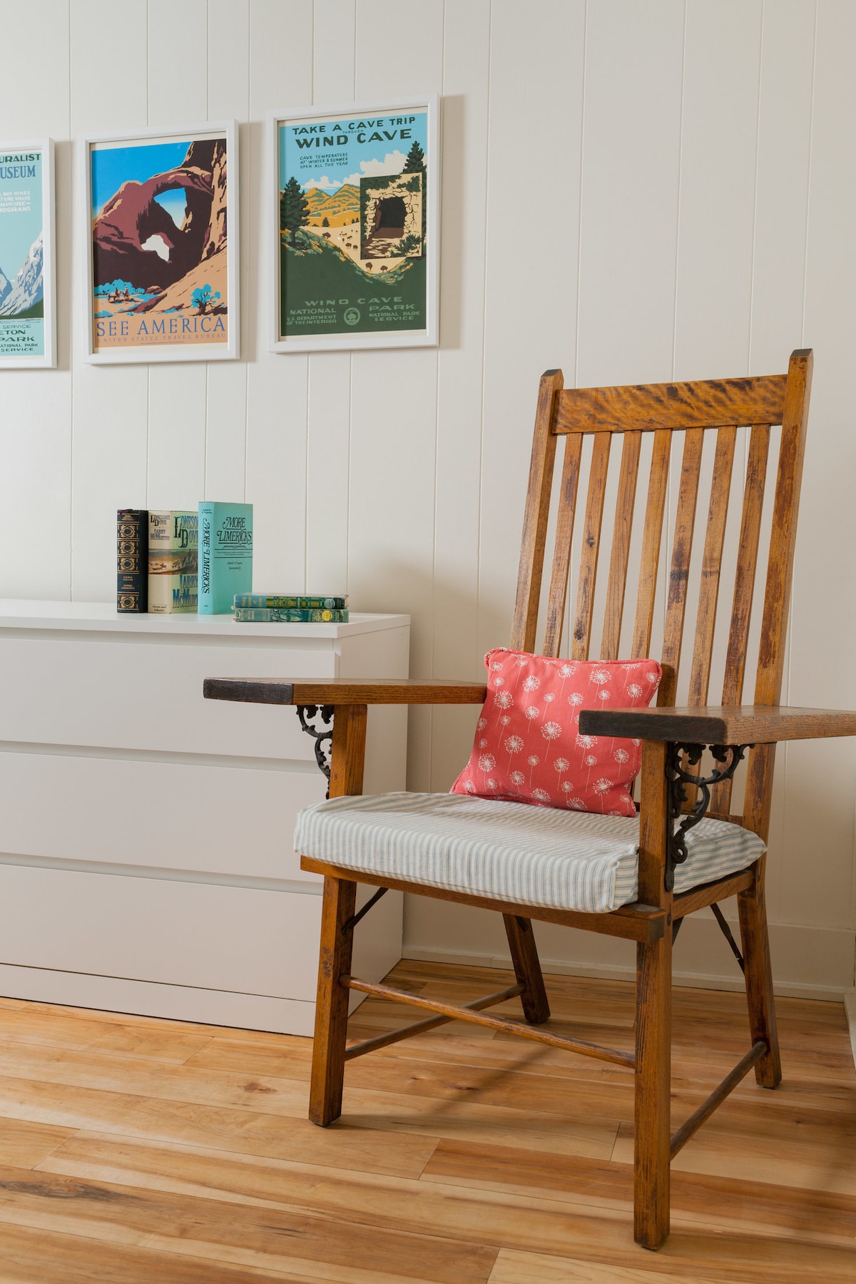 A wooden rocking chair with a cushioned seat sits beside a white dresser. Above, framed vintage travel posters are arranged in a gallery-style layout, adding a touch of character to the space. Natural light highlights the wooden floor, creating a warm environment.