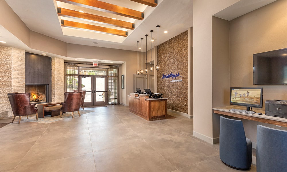 The spacious lobby features a reception area with a wooden desk and a cozy seating arrangement. Two chairs are positioned near a stone fireplace, offering a warm atmosphere. Large windows allow natural light to fill the space, complementing modern design elements.