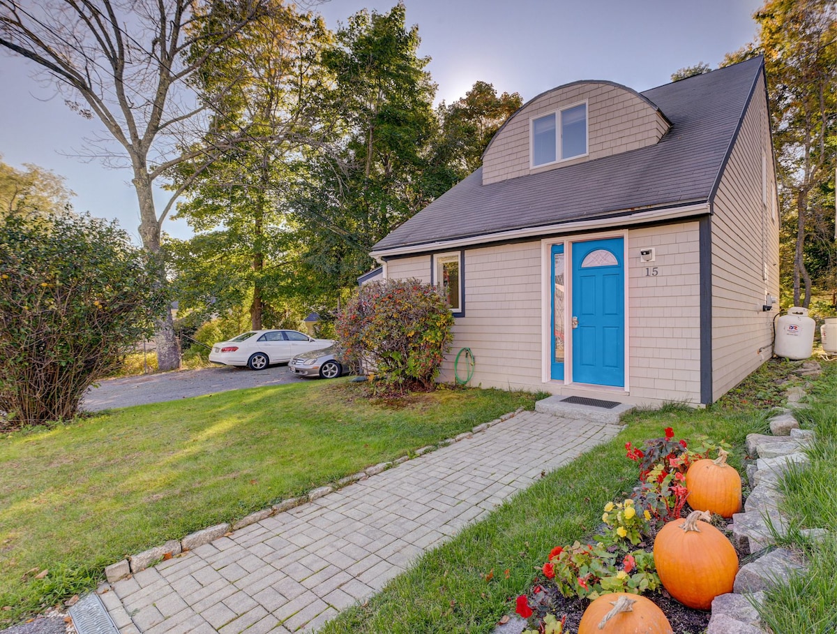The exterior of the house features a charming façade with a bright blue front door. A paved pathway leads to the entrance, bordered by well-maintained greenery and flower beds. Two cars are parked in the driveway, surrounded by autumn decorations, including pumpkins.