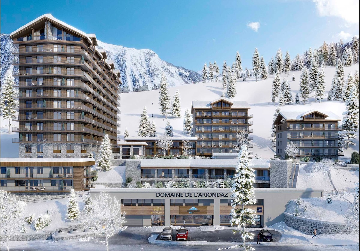 The image showcases a modern alpine resort surrounded by snow-covered mountains. Three distinct buildings exhibit contemporary architecture, with expansive balconies and large windows. Pine trees adorn the landscape, creating a serene winter atmosphere. The lower level features an entrance with signage for 'Domaine de L'Ariondaz.'