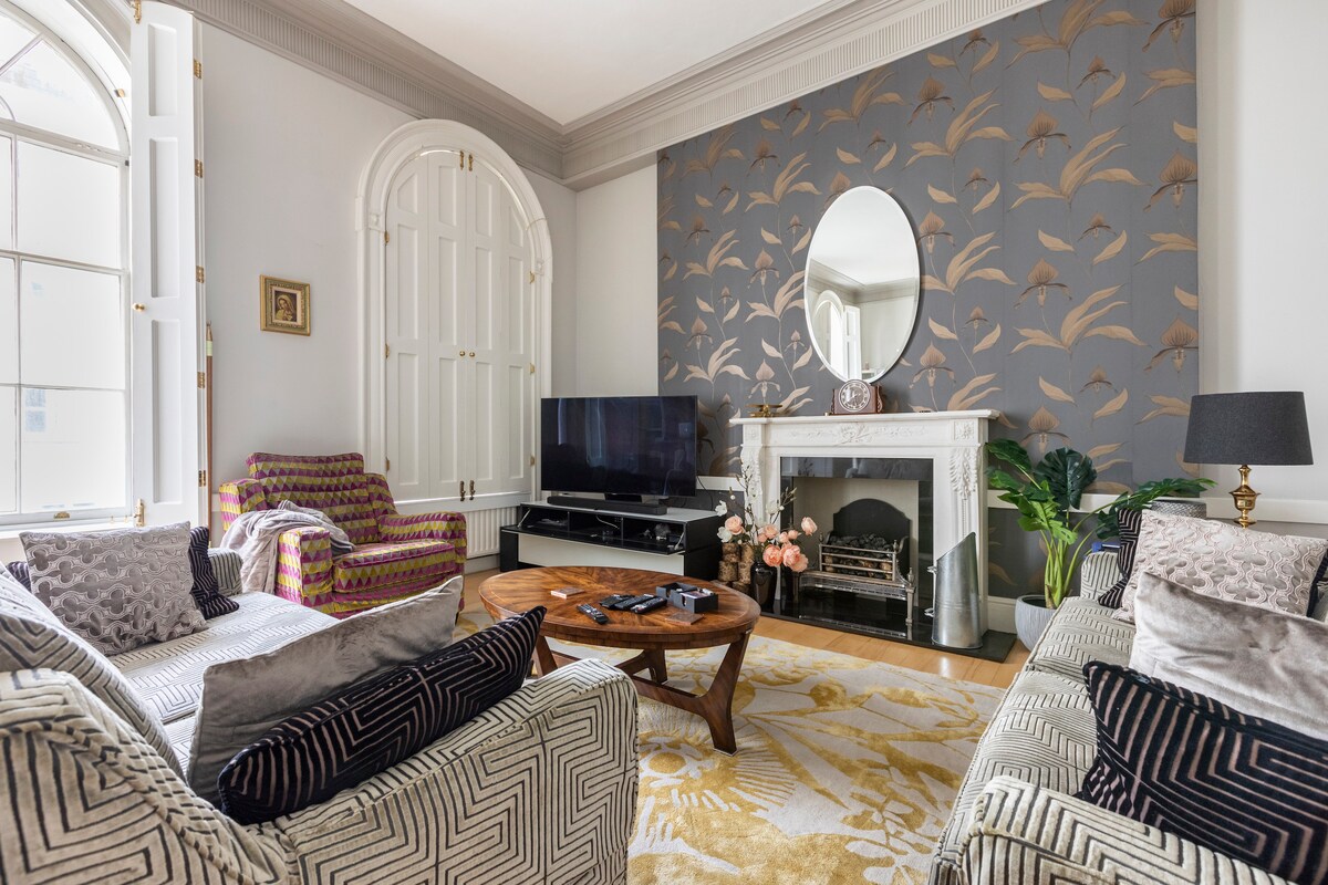 The living area features a stylish design with a striking patterned feature wall. A large mirror is positioned above the fireplace, reflecting light into the room. Two armchairs are arranged with a comfortable sofa, and a wooden coffee table is centered on a decorative rug.
