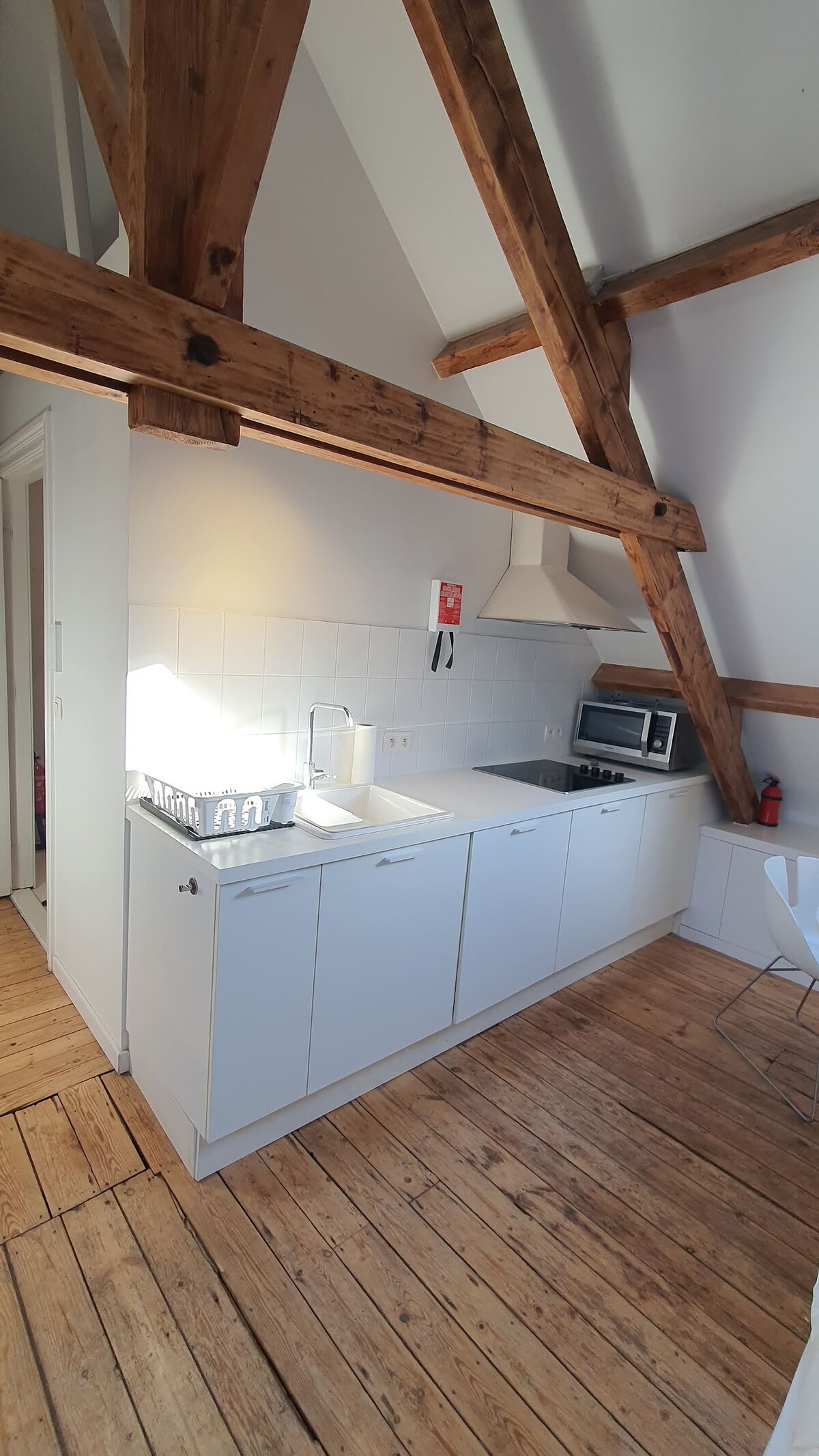 A well-equipped kitchen features modern appliances, including a stove, microwave, and a sink. Exposed wooden beams add character to the white cabinetry and tile backsplash. Natural light illuminates the space, highlighting the warm wooden flooring.