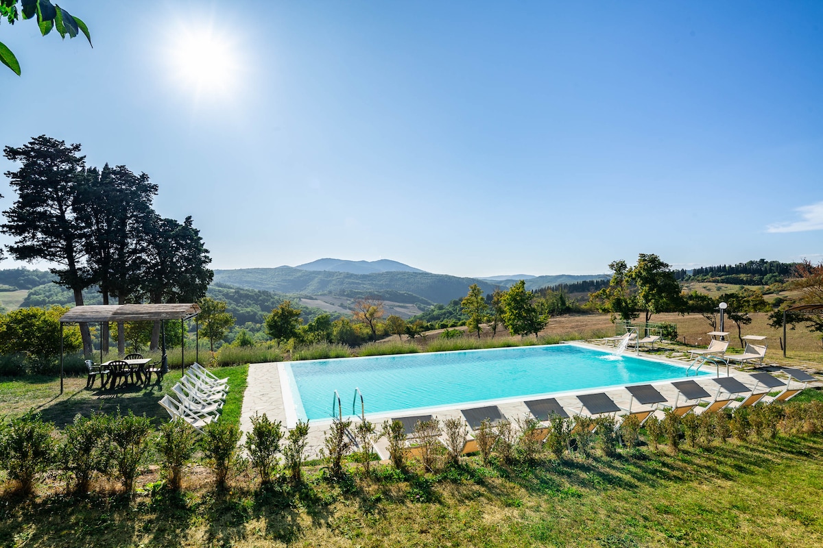 A swimming pool is set against a backdrop of rolling hills, with sunlight reflecting off the clear water. Sun loungers line the poolside, while shaded seating is provided under a pergola. Lush greenery and trees surround the pool area, enhancing the serene landscape.