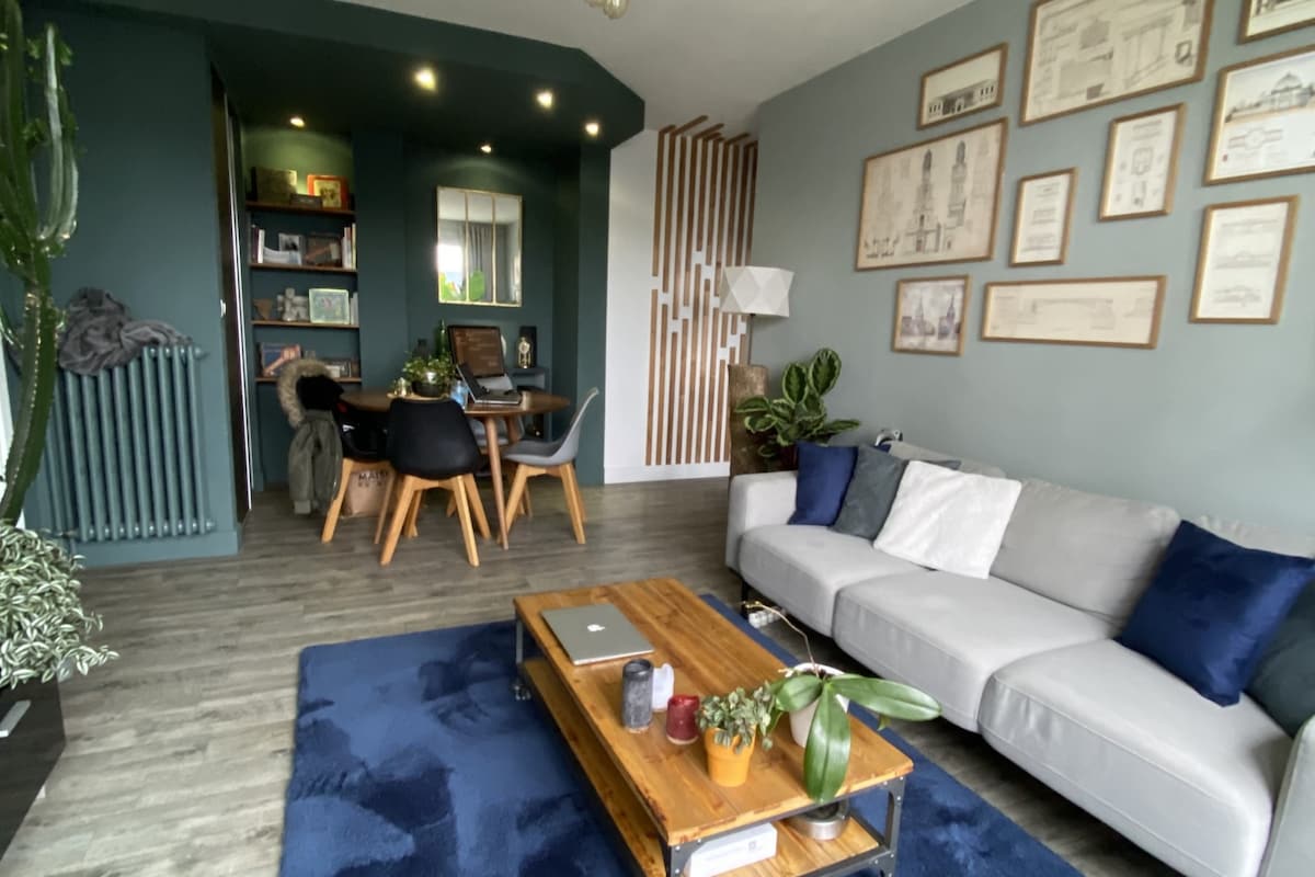 A modern living area features a cozy sofa with decorative pillows and a wooden coffee table. A dining space with stylish chairs is visible in the background, accompanied by bookshelves and greenery. Soft lighting and a textured wall enhance the inviting feel of the room.