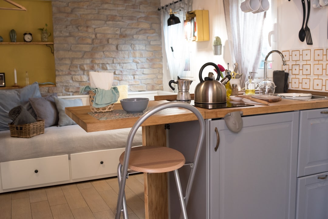 A well-equipped kitchenette features a wooden countertop and a modern kettle. Soft light highlights the stone wall and the cozy seating area in the background, which is adorned with cushions and a small decorative shelf.