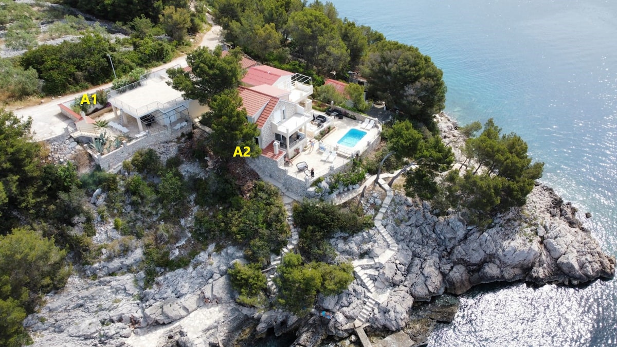 An aerial view of SESULA BAY RESORT offers a glimpse of two properties, A1 and A2, nestled among trees along the rocky coastline. A2 features a swimming pool, surrounded by outdoor furniture, with the shimmering sea visible in the background.